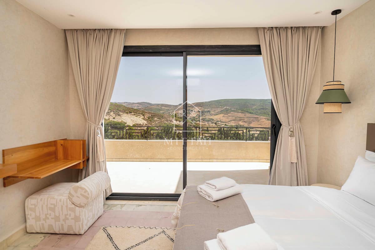 Habitación de hotel con cama, un banco de madera, un pequeño sofá y vistas del paisaje a través de un ventanal grande.