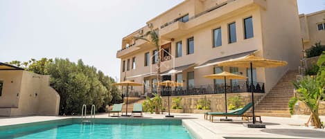 Uma área de piscina com espreguiçadeiras e guarda-sóis, um edifício com varandas e uma escadaria que leva aos andares superiores.