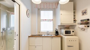 Cocina pequeña con lavamanos, microondas y refrigerador. Hay una ventana con cortina estampada, y un reloj en la pared.