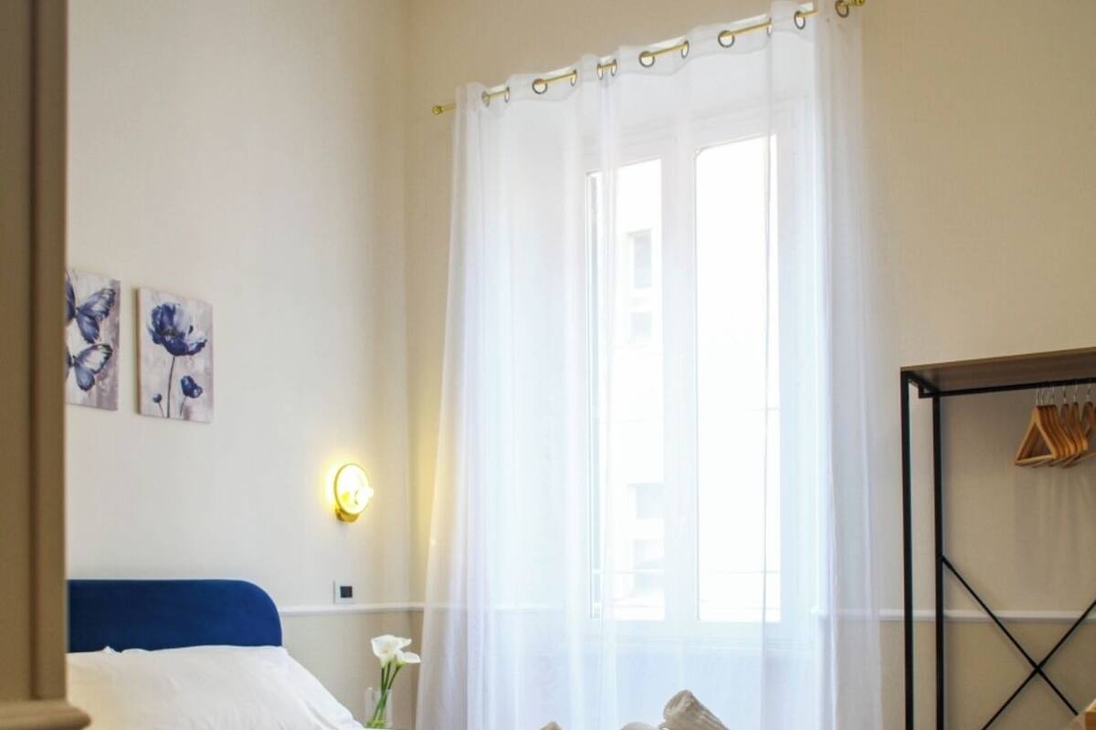 A bedroom with a bed, a bedside table with a lamp, a window with sheer curtains, and a wall-mounted coat rack.
