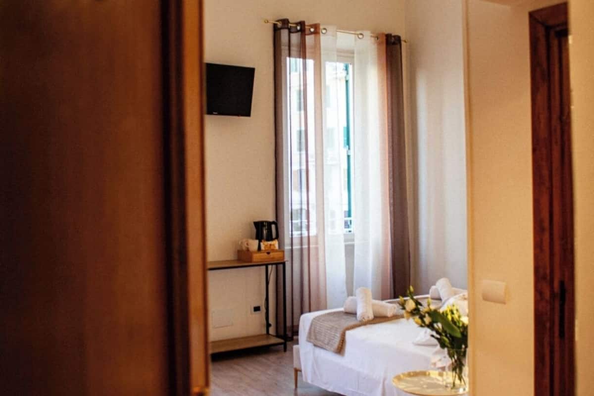 A hotel room with a wooden floor, a bed with white linens, a bedside table with a lamp, and a window with curtains.