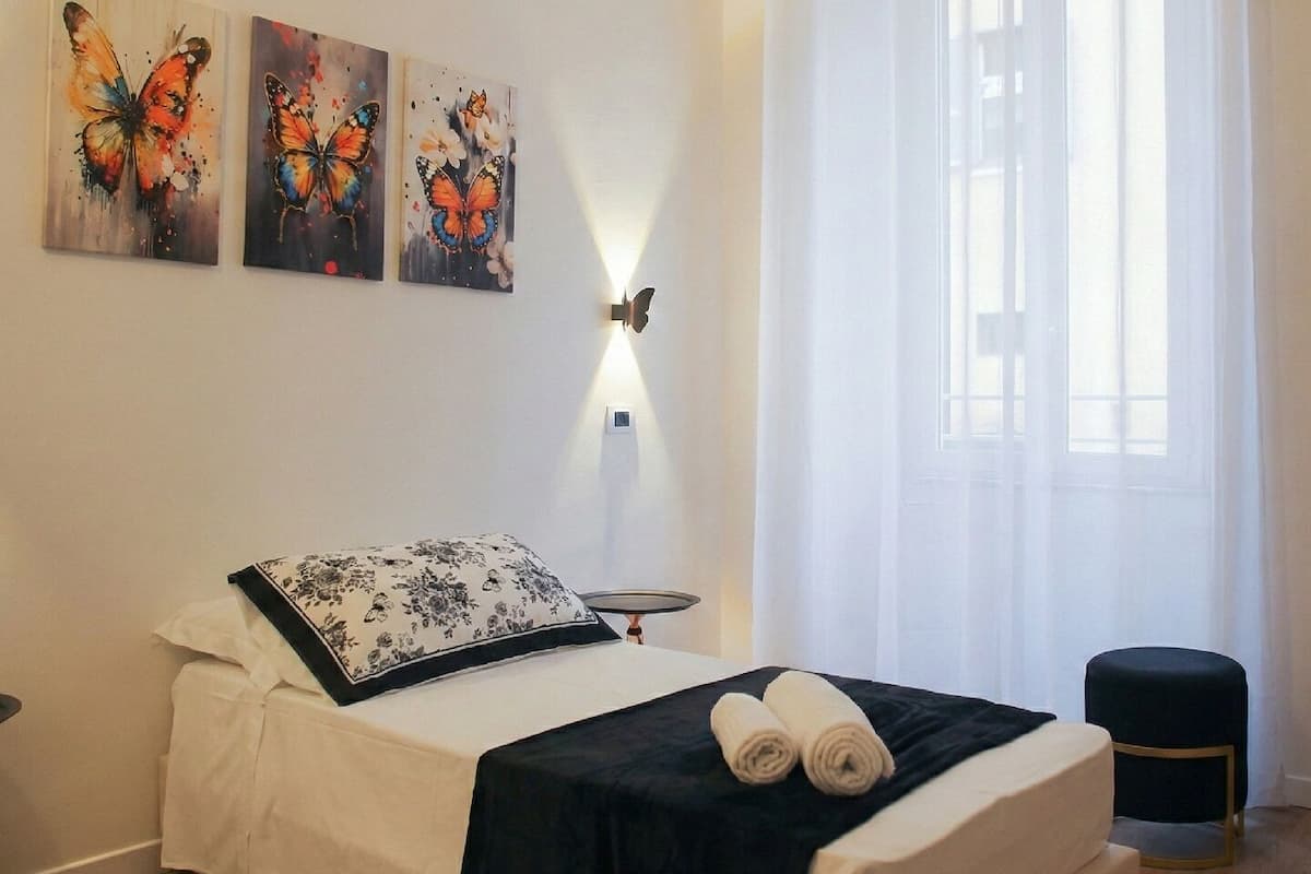 A bedroom with a bed, a bedside table, a lamp, a window with curtains, and three butterfly paintings on the wall.