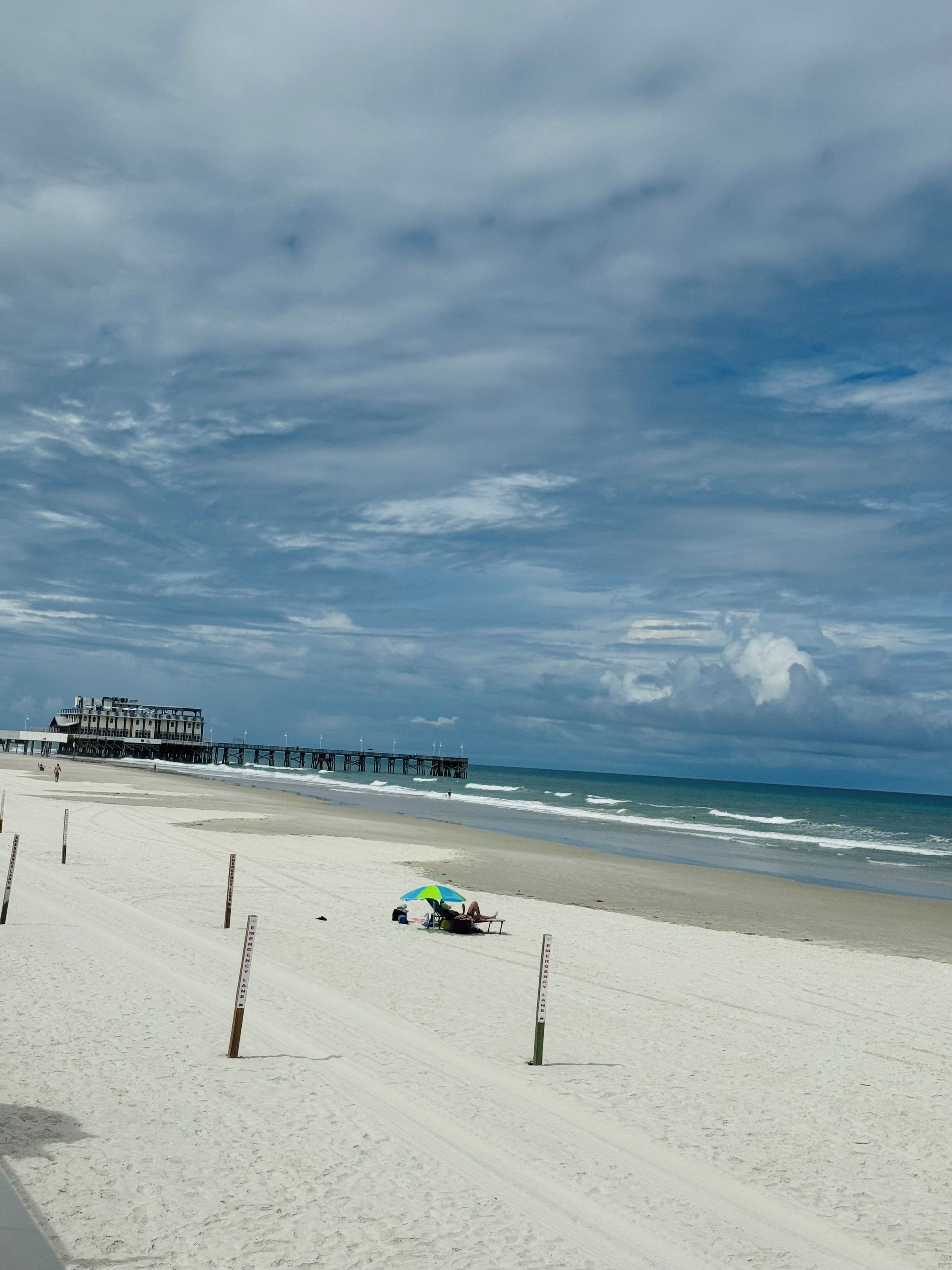 A beach with a pier, clear skies, and a person relaxing under an umbrella.