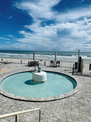 A pool with a dolphin statue, surrounded by a paved deck and a railing, overlooking the ocean. - Beach Vibes Only (Daytona Beach)