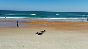 Una playa con cielo despejado, mar tranquilo y algunas personas caminando.