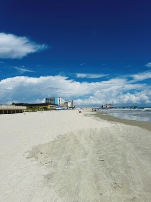Une plage avec une jetée qui s’avance dans l’eau, des bâtiments en arrière-plan et un ciel dégagé.