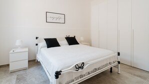 Een wit bed met zwarte accenten, een nachtkastje met een lamp, een lijst met een bord en een garderobe.