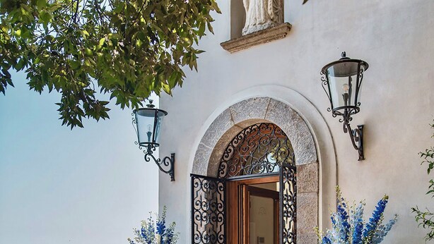 Une entrée en arc surmontée d’une façade en pierre, deux plantes en pot ornées de fleurs bleues et une vue sur la mer.