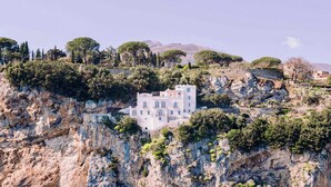 Un bâtiment blanc doté de nombreuses fenêtres, situé sur une falaise rocheuse, entouré d’arbres.