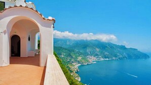 Un bâtiment blanc doté d’un sol carrelé et d’une porte en arc de cercle. Il offre une vue sur une zone côtière, avec la mer et les montagnes en toile de fond.