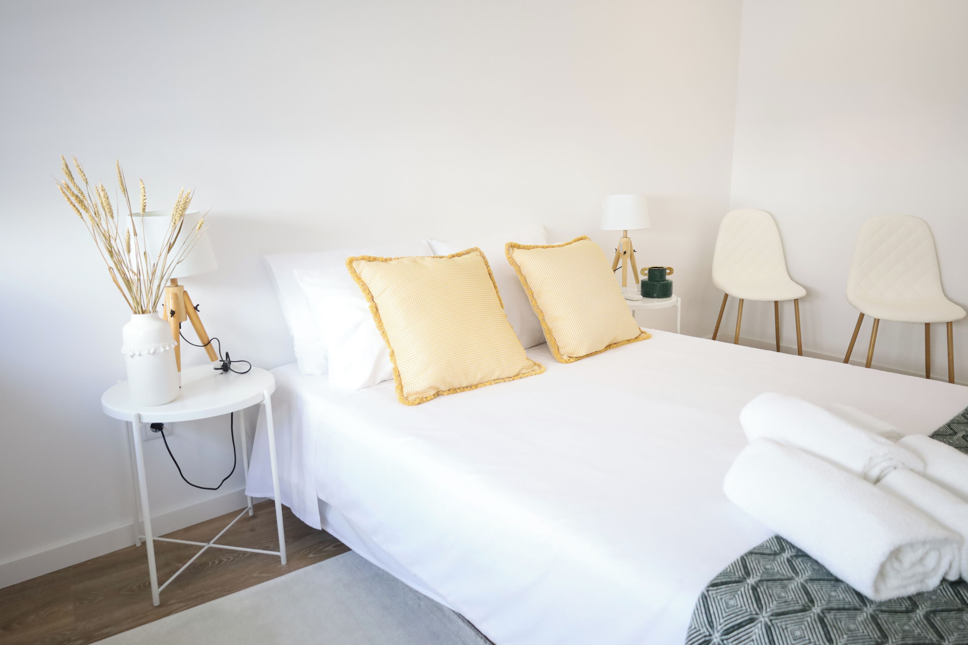 Une chambre d’hôtel bien rangée, avec un lit, deux oreillers, une table de chevet avec une lampe, un vase contenant des plantes séchées et deux chaises.