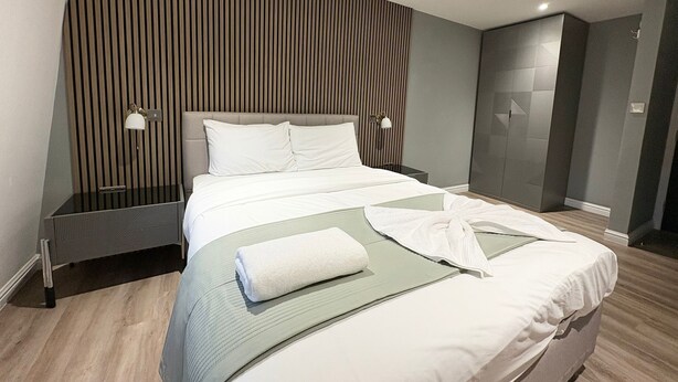 Una habitación de hotel moderna con una cama grande, dos lámparas de noche, un panel de pared de madera y un vestidor.