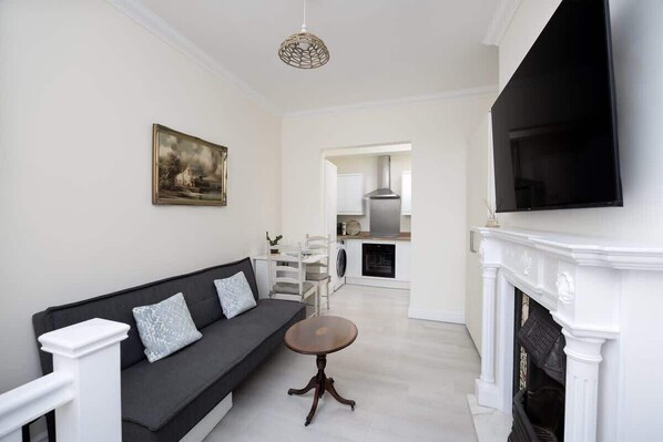 A living room with a sofa, a round table, a fireplace, a TV, and a kitchen in the background.