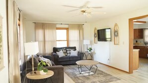 A living room with a sofa, two side tables, a ceiling fan, a TV, and a kitchen area visible through an open doorway.
