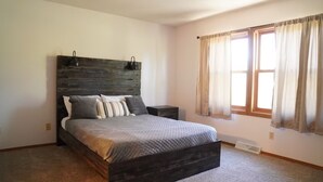 A bedroom with a wooden headboard, a bed with a gray quilt, a nightstand, and a window with curtains.
