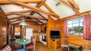 A cozy cabin-style room with wooden walls and ceiling, a dining table with chairs, a television on a wooden stand, and a window with curtains.