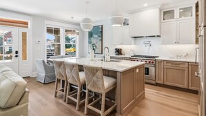 Eine moderne Küche mit einer zentralen Kochinsel, Barhocker, einem Essbereich mit einer Couch und Blick ins Freie.