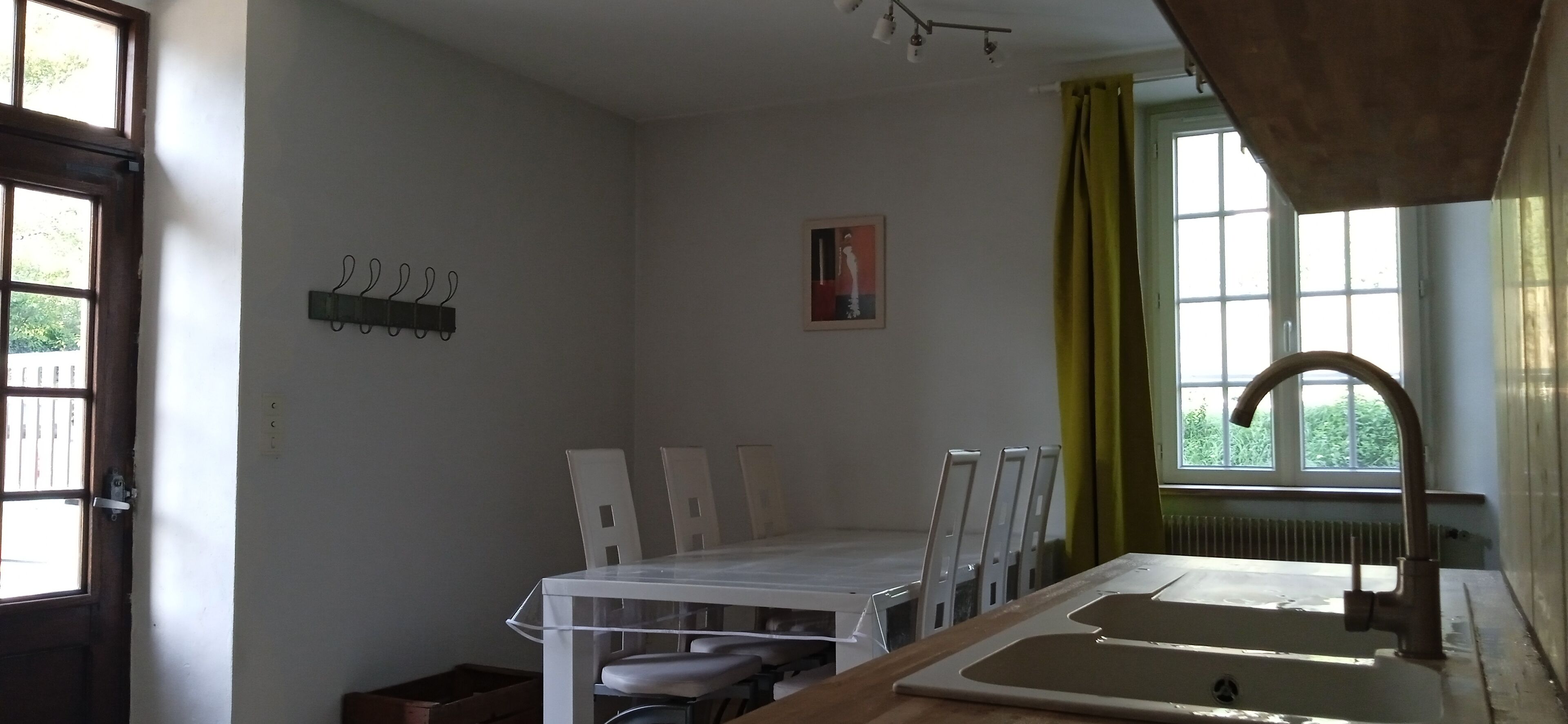 Una habitación pequeña y luminosa con una mesa de comedor y sillas, una ventana con cortinas, un radiador y un perchero en la pared.