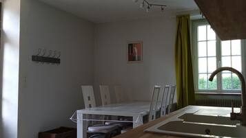 Una habitación pequeña y luminosa con una mesa de comedor y sillas, una ventana con cortinas, un radiador y un perchero en la pared.