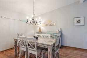 Ein Essbereich mit einem rustikalen Holztisch und Stühlen, einer Deckenleuchte, einem an der Wand befestigten Schild mit der Aufschrift 'BEACH HOUSE' und einer dekorativen Lampe.