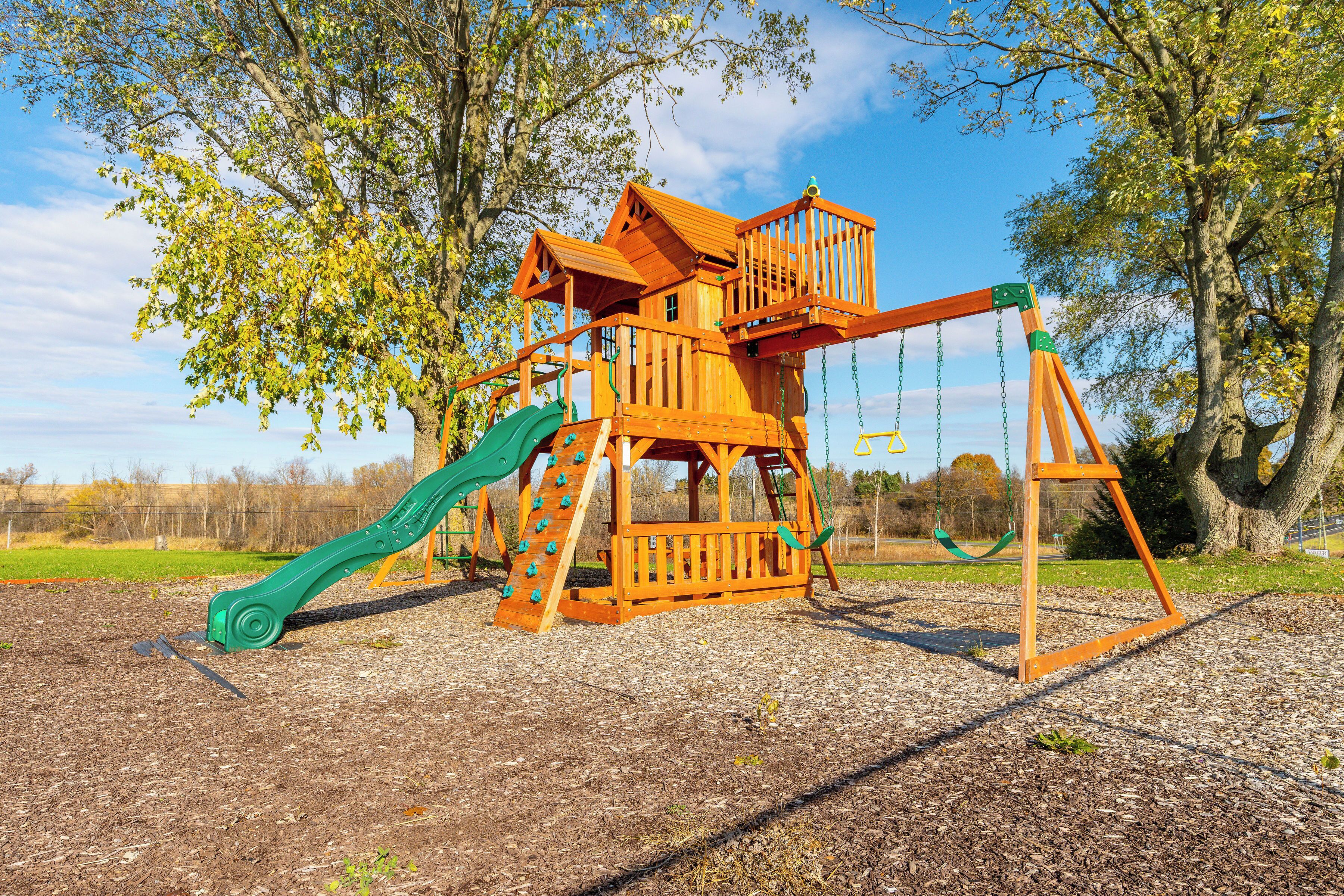 A wooden playground structure with a slide and swings, surrounded by trees and a clear sky.