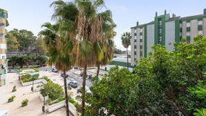 Un immeuble à plusieurs étages doté d’une façade verdoyante, un palmier au premier plan, et une zone pavée agrémentée de jardinières et d’une rue.