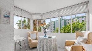 Une pièce d’angle avec un mur en briques blanches, une chaise blanche, une table ronde recouverte d’une nappe à motifs de feuilles, et une vue sur la verdure et les bâtiments environnants.