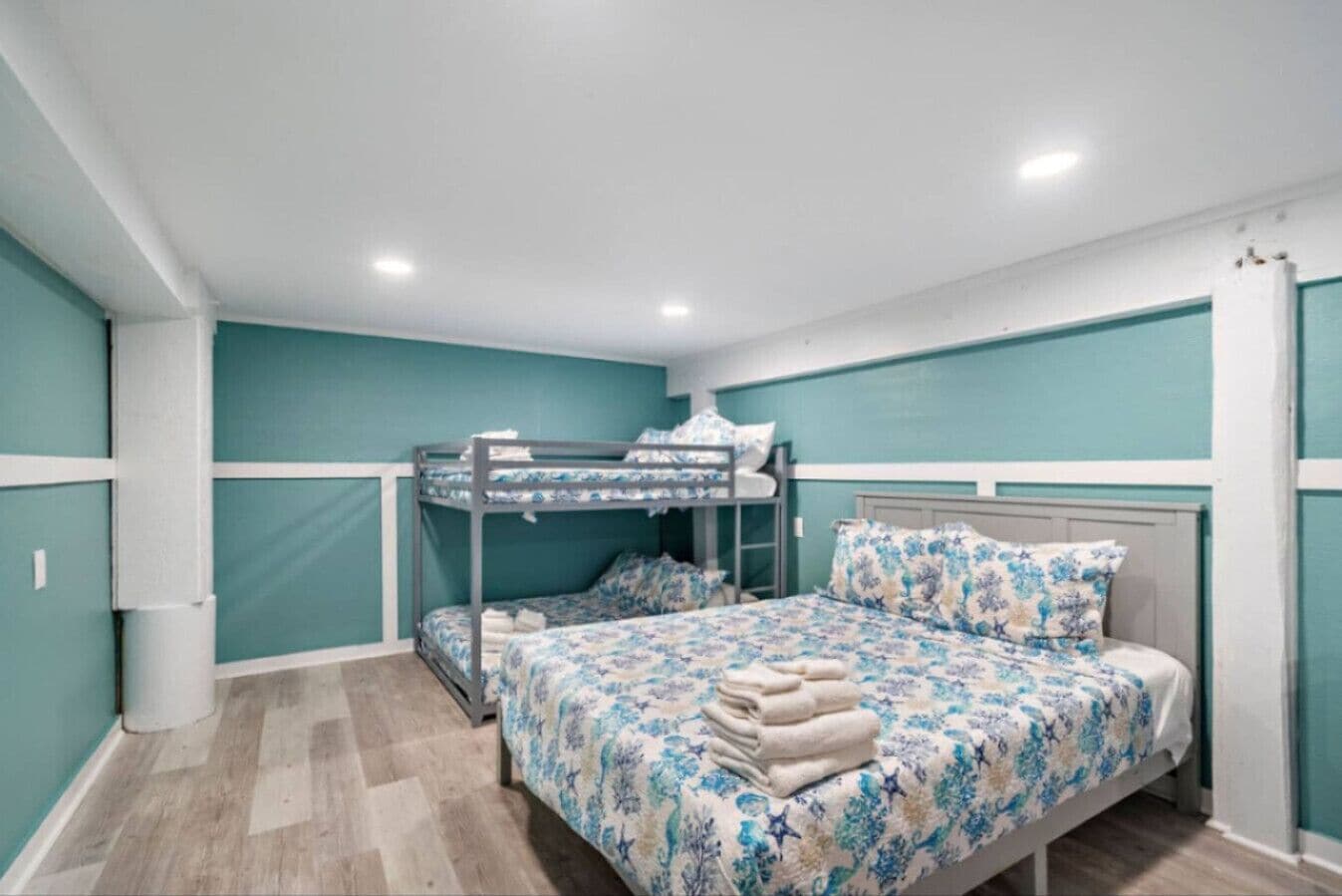 Una habitación con paredes color turquesa, una litera, una cama individual con ropa de cama floral y suelo de madera.