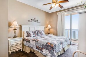 A bedroom with a bed, two bedside tables, lamps, a ceiling fan, a decorative wall piece, and a balcony with curtains. - Sterling Reef 405 (Panama City Beach)