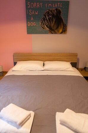 Una cama bien hecha con sábanas blancas y dos toallas dobladas al pie. Un cartel en la pared sobre la cama dice 'SORRY I'M LATE, I SAW A DOG'.