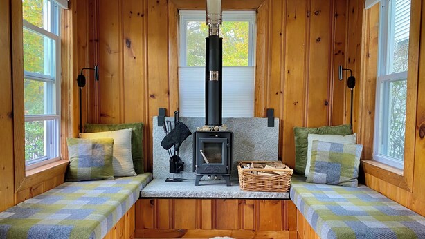 A cozy cabin interior with wooden walls, a stone hearth, a black wood stove, and two bench seats with checkered blankets. - Stay at adorable mill house in walkable Maynard. Small but well-designed spaces. (Maynard)