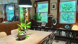 A wooden dining table with a bowl of fruit and a potted plant, a bookshelf with books, and a window with a view of greenery. - Stay at adorable mill house in walkable Maynard. Small but well-designed spaces. (Maynard)