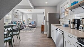 A modern kitchen with stainless steel appliances, a dining area with a wooden table and chairs, and a living room with a sofa and a coffee table. - Charming Home in Perfect Central Spot (Galveston)