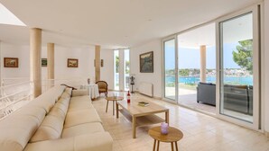 A spacious living room with a large sofa, a dining table, and a view of the sea. - Villa Ses Arcades II by Villa Plus (Cala d'Or)