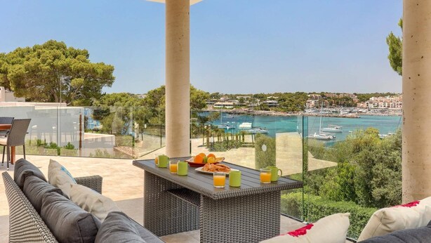 A balcony with wicker furniture, a glass table with breakfast items, and a view of a coastal town with boats. - Villa Ses Arcades II by Villa Plus (Cala d'Or)