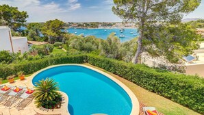 A swimming pool with lounge chairs, surrounded by greenery and overlooking a coastal area with boats. - Villa Ses Arcades II by Villa Plus (Cala d'Or)