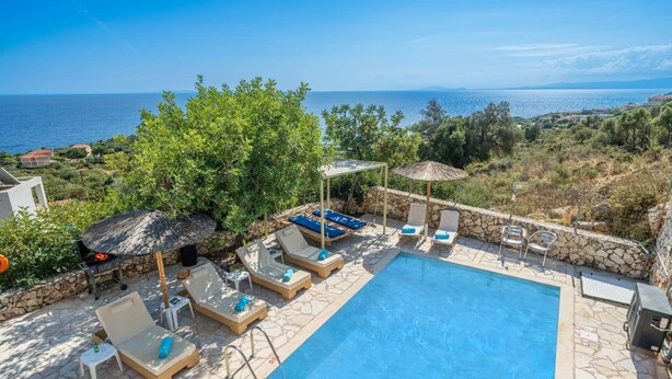 A pool area with lounge chairs and umbrellas, surrounded by stone walls and vegetation, overlooking a body of water. - Villa Apollon by Villa Plus (Ionian Islands)