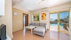 A modern living room with a grey sofa, a small white table, and a TV. There is a glass door leading to a pool area with a view of the outdoors. - Villa Apollon by Villa Plus (Ionian Islands)