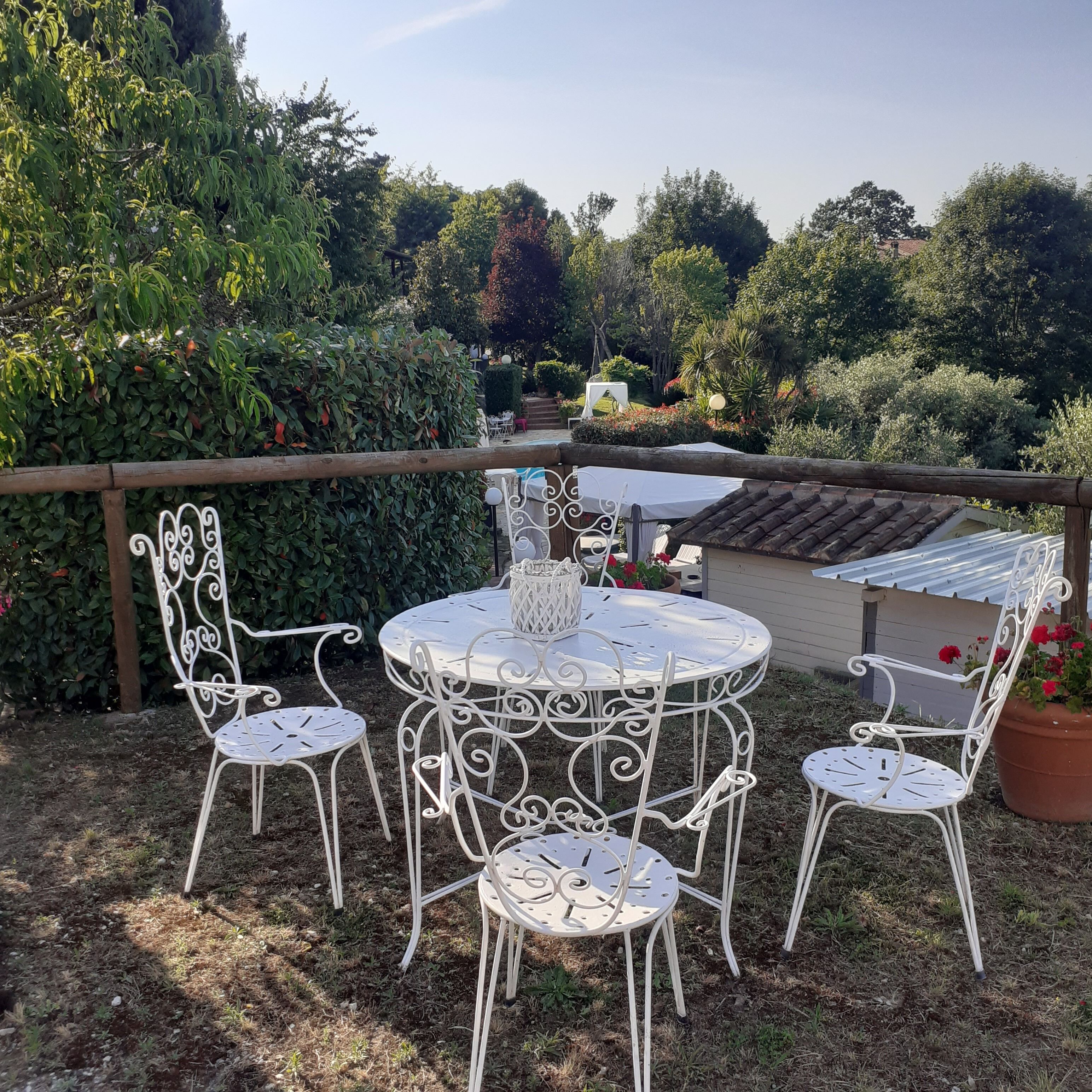 An outdoor patio with a round metal table and white metal chairs, surrounded by lush greenery and a wooden fence.