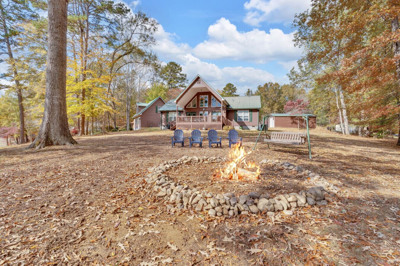 A cabin with a stone fire pit, surrounded by autumn leaves, and blue Adirondack chairs.