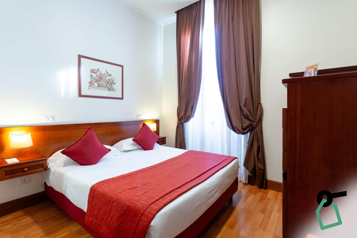 A hotel room with a wooden bed, red bedding, a nightstand with a lamp, a framed floral artwork on the wall, and a wooden wardrobe.