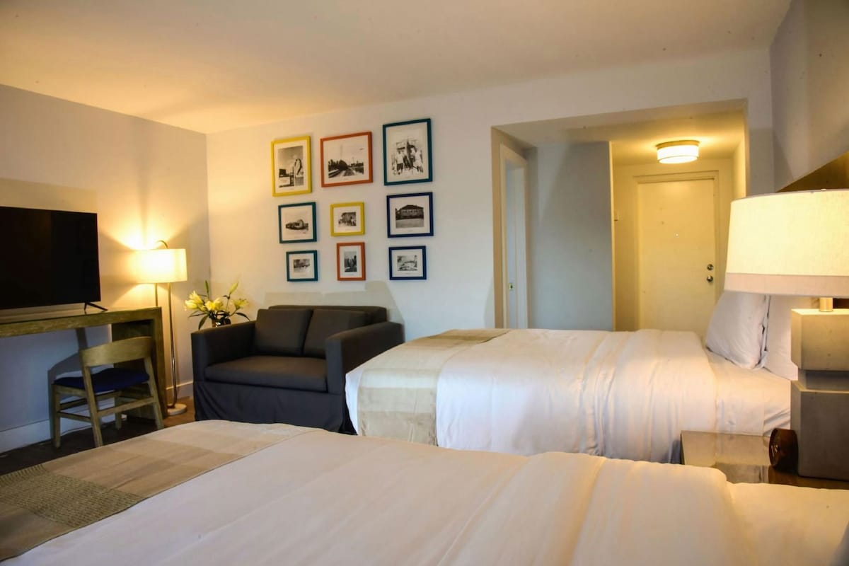 Ein Hotelzimmer mit zwei Betten, einem Fernseher, einem Schreibtisch, einem Sessel und gerahmten Bildern an der Wand.