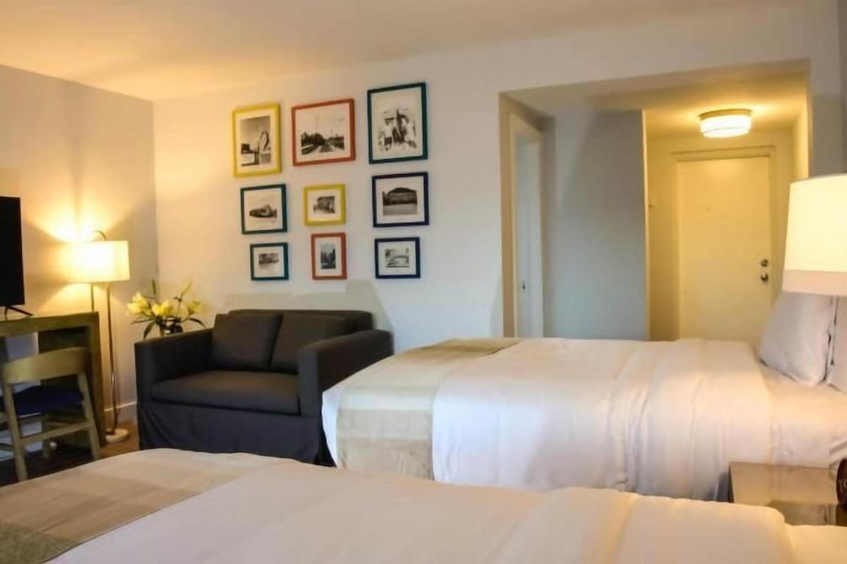 Ein Hotelzimmer mit zwei Betten, einem Fernseher, einem Schreibtisch und gerahmten Bildern an der Wand.