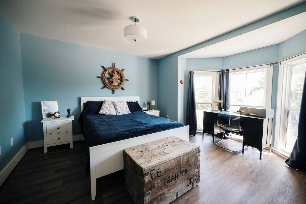 Ein Schlafzimmer mit Bett, Schreibtisch, Stuhl, einer Holzkiste, einem Nachttisch, einer Wanduhr und einer Schiffslenken-Dekoration.