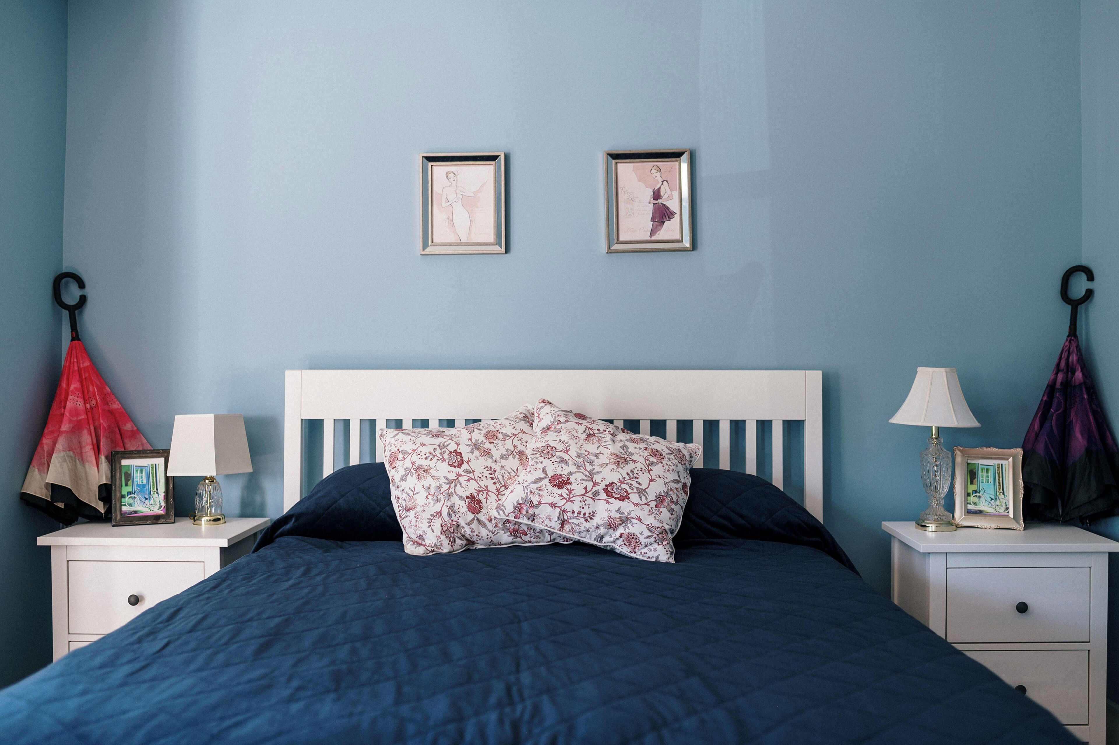 A bedroom with a bed, two framed pictures on the wall, a nightstand with a lamp, and a coat hanging on a hook.