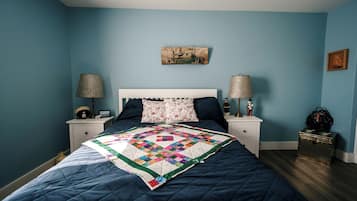 Un dormitorio con pared azul, cabecera blanca, colcha, dos mesitas de noche con lámparas y una maleta en el suelo.