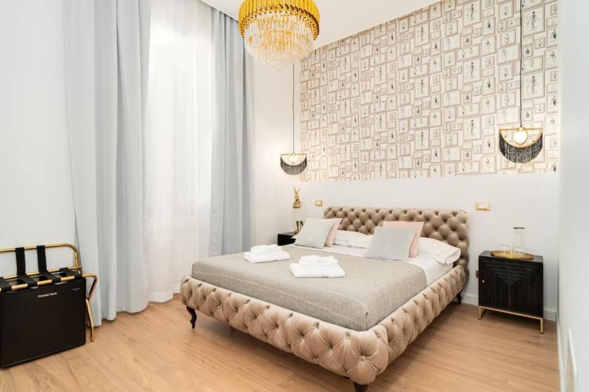 Un dormitorio con un cabecero capitonado, una cama grande con toallas blancas ordenadas, una pared con cuadros enmarcados y una mesita de noche negra con una lámpara.
