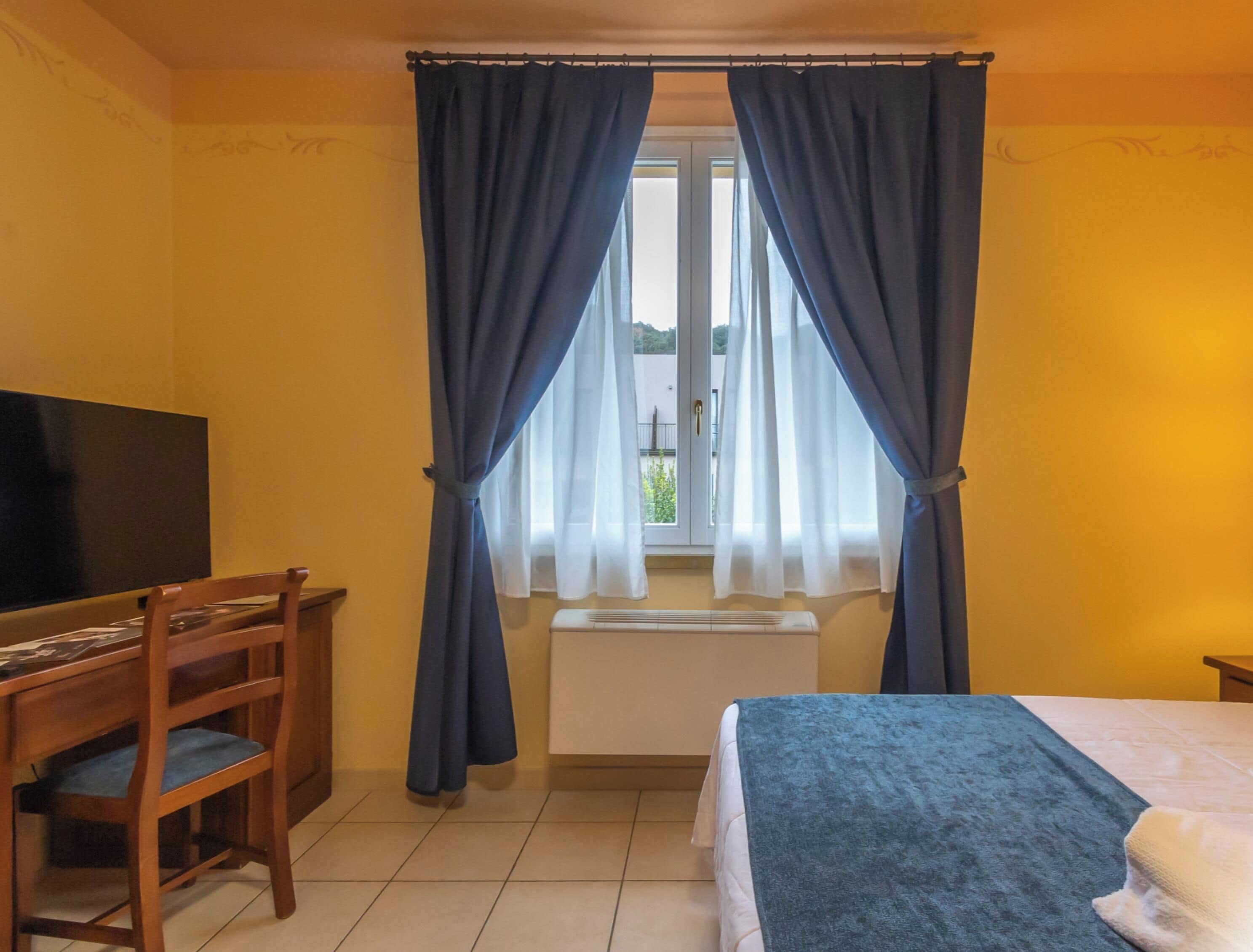Una camera d'albergo con un letto, una scrivania con una sedia, una televisione e una finestra con tende.