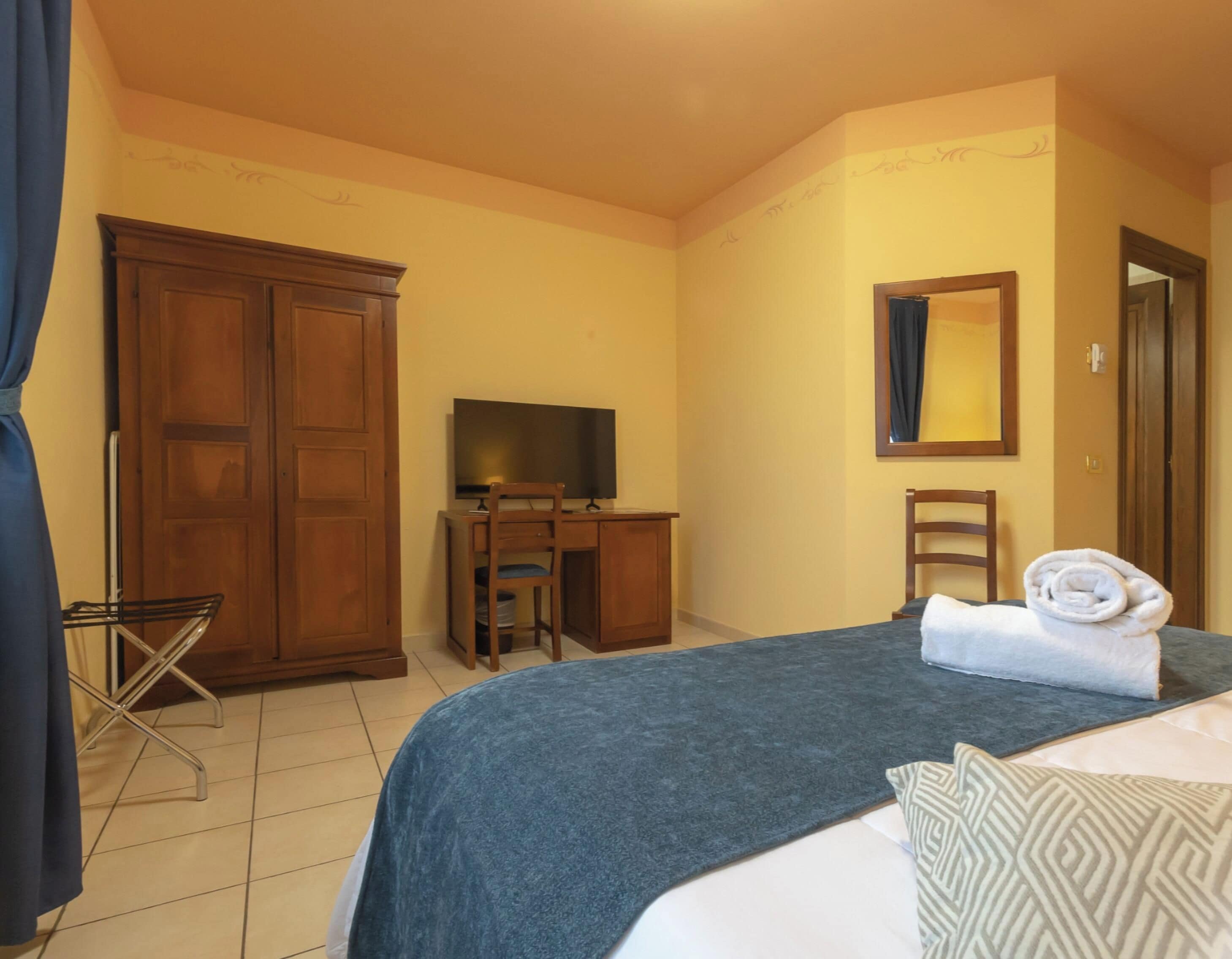 A hotel room with a bed, a desk with a computer, a chair, a wardrobe, and a mirror.