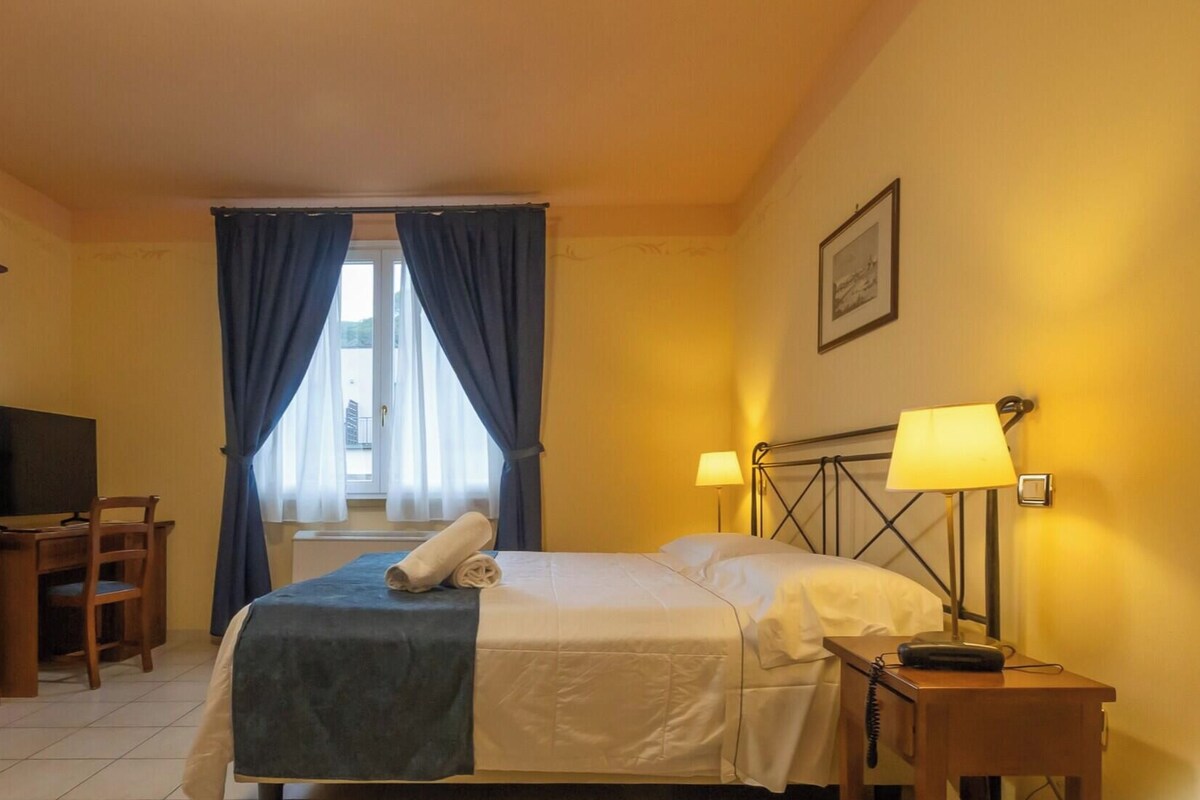 A hotel room with a bed, bedside lamps, a desk with a chair, a television, and a wardrobe.